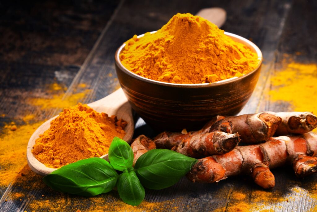 Bowl and spoon overflowing with powdered tumeric next to its roots on wooden surface